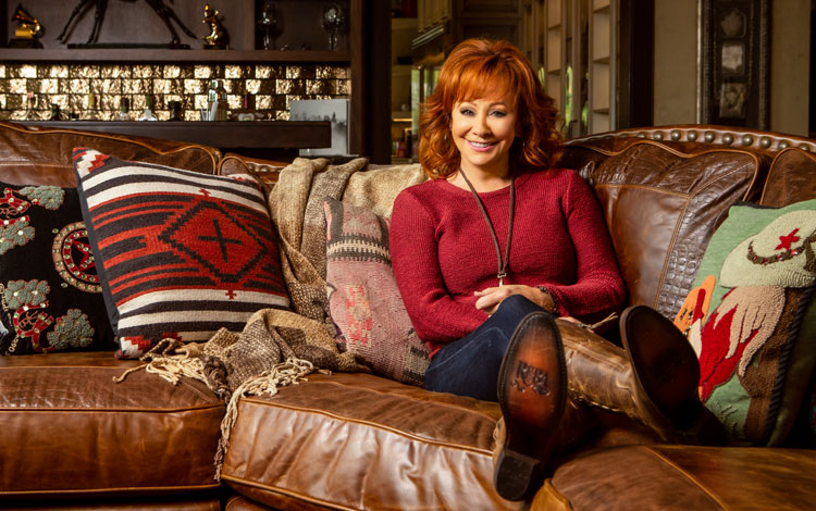 Reba McEntire with fiery red hair wearing a red long-sleeve shirt, sitting on a couch